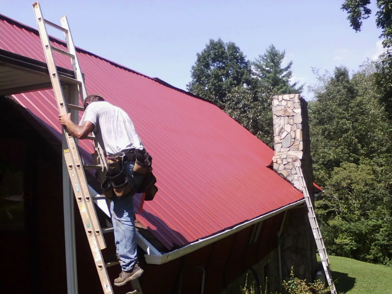 Roofer on a ladder beside a red metal roof and stone chimney — Soffit & Fascia Repair by Roofing Brothers in Accord
